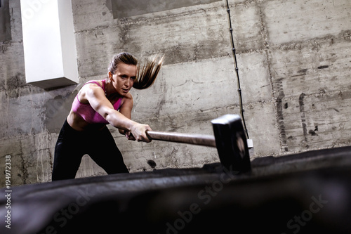 Fotomural Fitness woman hitting wheel tire with hammer sledge in the gym, cross fit training
