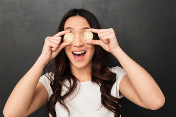 Wall Mural - Excited emotional asian young woman holding two golden bitcoins covering eyes.