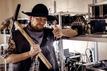 A man in a hat and with a tool on his shoulder shows a good gesture against the background of computer devices