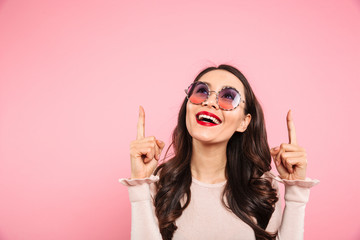 Poster - Content adult girl with red lips wearing round sunglasses looking upward and pointing fingers on copy space, isolated over pink background