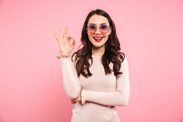 Wall Mural - Adorable woman 20s in trendy round glasses looking on camera and showing alright sign with fingers, isolated over pink background