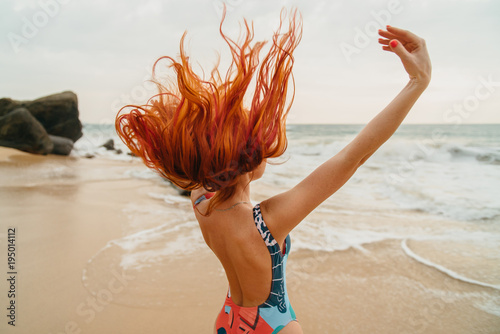 Photographie young red-haired woman with flying hair on the ocean coast at sunset, rear view