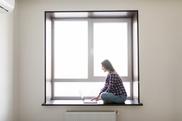 Beautiful young woman sitting on windowsill and using laptop against window