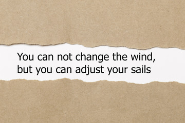 Motivational quote You can not change the wind but you can adjust your sails, appearing behind torn paper.