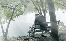 Lonely Man Sitting On Bench Under Tree In Park With Hopeless Feeling In Front Of Lake