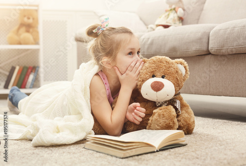 Fotomural Happy little female child and her teddy bear reading book on the floor at home