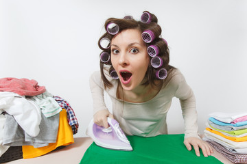 Wall Mural - Close up fun young housewife with curlers on hair in light clothes ironing clothing on ironing board with iron. Woman isolated on white background. Housekeeping concept. Copy space for advertisement.