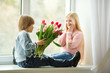 © _chupacabra_ - Pretty kids are sitting on window sill , happy little boy giving a cute girl bouquet of pink tulip flowers. Easter concept