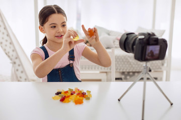 Love sweets. Pleasant cute pre-teen girl holding two gummy bears and reviewing them on camera while recording a video blog