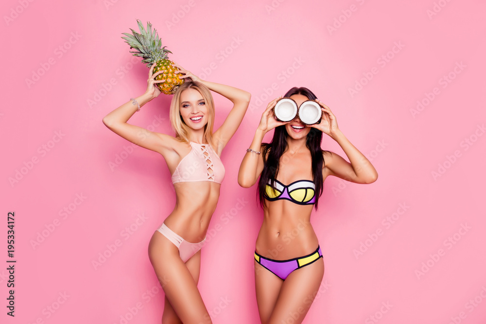 Portrait of hot, charming, stylish, pretty, trendy, crazy ladies, tourists in swim suits, blonde having ananas on her head, brunette having coconut on eyes' place, standing over pink background - obrazy, fototapety, plakaty 