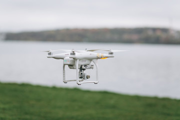 White quadrocopter is flying high in the air, taking photos, recording footage from above. Drone with four motors and propellers, camera and red warning lights.