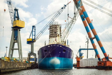 An old tanker moored in a dockyard, undergoing maintenance or repair, surrounded by large lifting cranes.