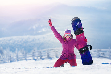 Snowboarding. Young woman sits on snow and holds snowboard, raises her hand upwards and smiles.