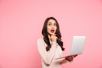 Wall Mural - Portrait of caucasian woman with long dark hair holding silver laptop and looking aside on copyspace with curiosity, isolated over pink background