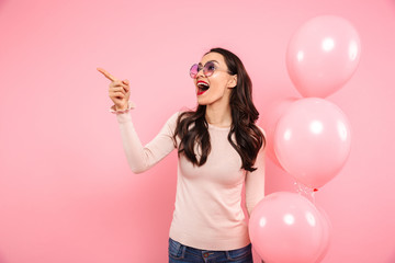 Wall Mural - Photo of delighted adult girl with long dark hair in round glasses holding lots of balloons and pointing finger aside, isolated over pink background