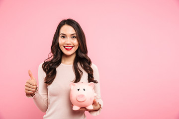 Wall Mural - Lovely satisfied woman 20s with long dark hair holding piggybank and gesturing thumb up, isolated over pink background