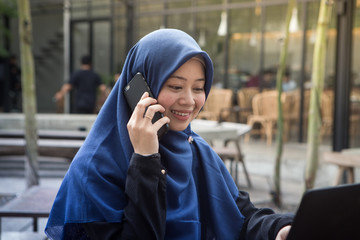 Beautiful southeast asian hijab woman working on laptop and answering phone at outdoors cafe