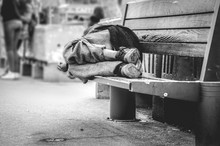 Poor Homeless Man Or Refugee Sleeping On The Wooden Bench On The Urban Street In The City, Social Documentary Concept, Selective Focus, Black And White