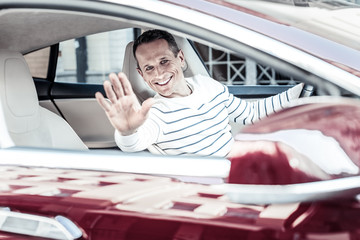 Car driver. Happy positive nice man sitting behind the wheel and waving his hand while greeting a friend