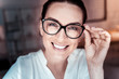 Good joke. Satisfied pleasant bespectacled woman looking straight supporting her glasses and smiling.