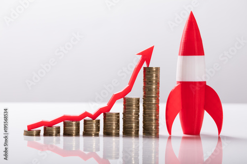Fotografie, Obraz Close-up Of Red Rocket Besides Stacked Coins And Arrow