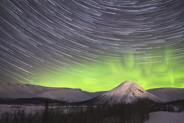 Wall Mural - Star Trails on Night Sky and Green Northern Lights in Snowy Mountains at Winter Night.