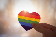 Lgbt Rights Concept, Hand Holds A Heart Painted Like A LGBT Flag, Silhouetted Against Sun