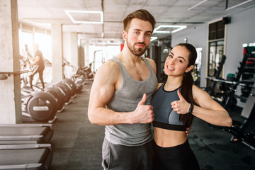 A picture of a couple standing in the sport club's training room together and showing their big thumb's up. They are happy to exsercise in this fitness club. Young couple is smiling.