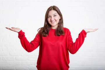 A young pretty smiling girl in a red sweater and a hat against a modern white brick wall in the loft style. She looks at the viewer. Copy space.