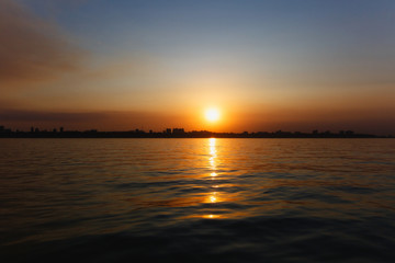  Beautiful sea landscape. Bright warm sunset in the sea near the coastline with the city's silhouettes. Sun hides behind the house. Colors of the sky are reflected in the water
