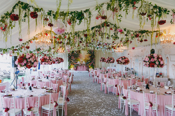 Beautiful decoration of the wedding banquet under the awning in pink, burgundy and white tones. Hall with dining tables and chairs decorated with thin cloth, bouquets, flower garlands and confetti