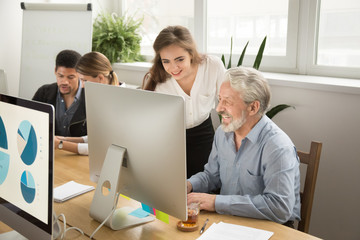 Smiling young manager helping senior worker with funny computer work in office, mentor teacher training happy older employee at workplace, colleagues of different age laughing looking at pc monitor