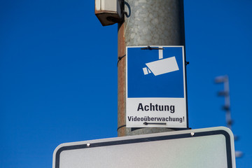 monitoring camera sign hangs on a lamp pole. Achtung Videoueberwachung means caution video control