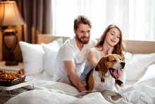 Funny Dog Yawns While Sits In Bed Near Hosts, Satisfied With Delicious Breakfast. Happy Female And Make In Background Take Care About Their Favourite Pet. People, Lifestyle And Rest Concept.