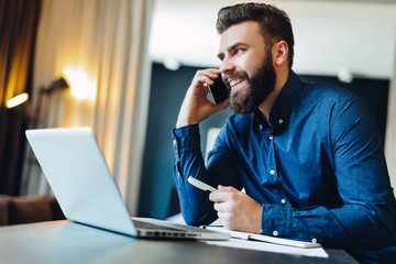 Young smiling bearded businessman is sitting at table in front of computer, talking on cell phone, holding pen. Freelancer works at home. Telephone conversations. Distance work, online education.