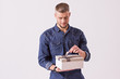 Studio shot of bearded hipster man tying gift box and smiling while isolated on white background. Young handsome man in denim shirt opening birthday present against white background.