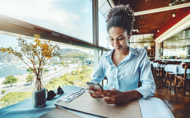 Young smiling lovely Brazilian girl is waiting for her food order while sitting next to the window in a luxury restaurant on the roof of a house and having online chat in smartphone with her friend