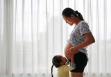 Fototapeta Sypialnia - Pregnant Asian woman standing near window at home with her daughter kissing her tummy.
