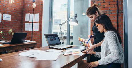 Young women working on a new web design using color swatches and sketches sitting at desk in modern office