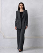 Gorgeous young woman dressed in gray squared jumpsuit, blazer and heeled shoes posing in studio. Beautiful brunette girl demonstrating stylish smart clothing against white wall on background.