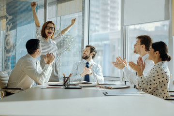 Perfect day. Adorable young woman raising her hands in joy and celebrating her promotion while her colleagues clapping their hands and congratulating her