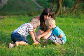 Mother with dark hair is playing with two children in the green lawn. Happy fun summer photo. Smiling laughing boys with his mom. Sunny day outdoors in nature. Kids and mom are hugging.