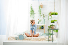 Young Woman Having A Good Morning Sitting On The Bed In The Beautiful Bright Bedroom With Green Plants And Clock