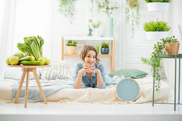 Wall Mural - Portrait of a young and beautiful woman lying in the bright and cozy bedroom with fresh green food and plants at home