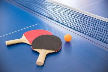 Orange ball for table tennis and two rackets of red and black color on a blue table with a grid