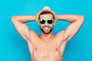 Attractive, cheerful, manly, stunning, funny man in glasses and hat holding hands behind the head, showing his shaven armpits on holidays, enjoying sunshine, taking sunbath over blue background