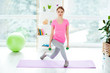 Front view portrait of powerful active focused beautiful energetic attractive woman holding green dumbbells in hands doing sit-ups standing on purple mat training in light modern white studio club gym