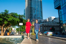 Portrait Of Couple In Bright Clothes And Sunglasses Holding Hands, Laughing During Walk In The Street With Skyscrapers. Happiness, Lifestyle And Tourism Concepts. Selective Focus. Copy Space.