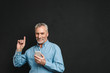 © Drobot Dean - Image of bearded male pensioner 60s with gray hair pointing finger upward like have idea while using mobile phone, isolated over black background