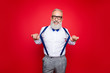 © deagreez - Portrait of elegant, confident, playful macho, old barber, stylist draw off suspenders with thumb fingers, looking at camera with beaming smile, isolated on red background